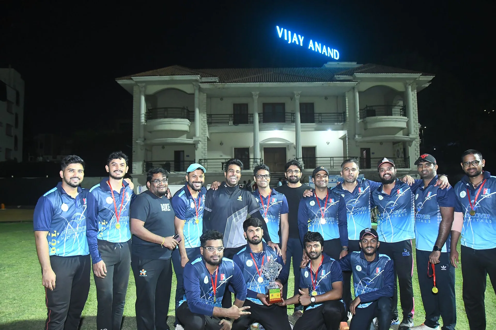A Doctors Cricket League team in blue kit posing together on the ground under floodlights